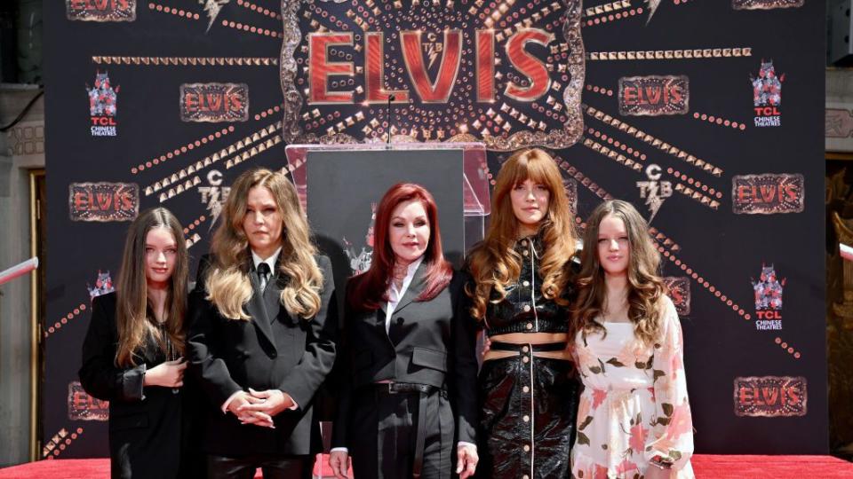 harper lockwood, lisa marie presley, priscilla presley, riley keough, and finley lookwood stand together for a photo in front of a stage and a backdrop that says elvis