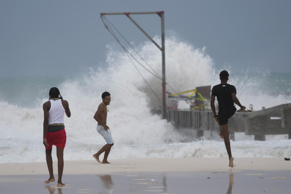 Three youths near the ocean watch a large wave crashing onto the beach.