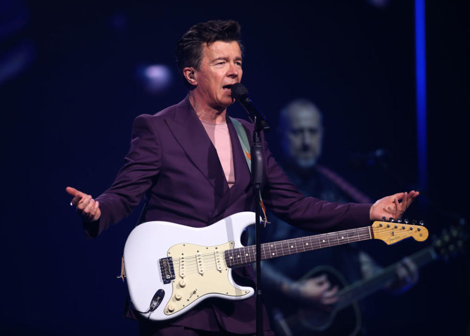 Rick Astley performs on stage holding a guitar, wearing a tailored suit