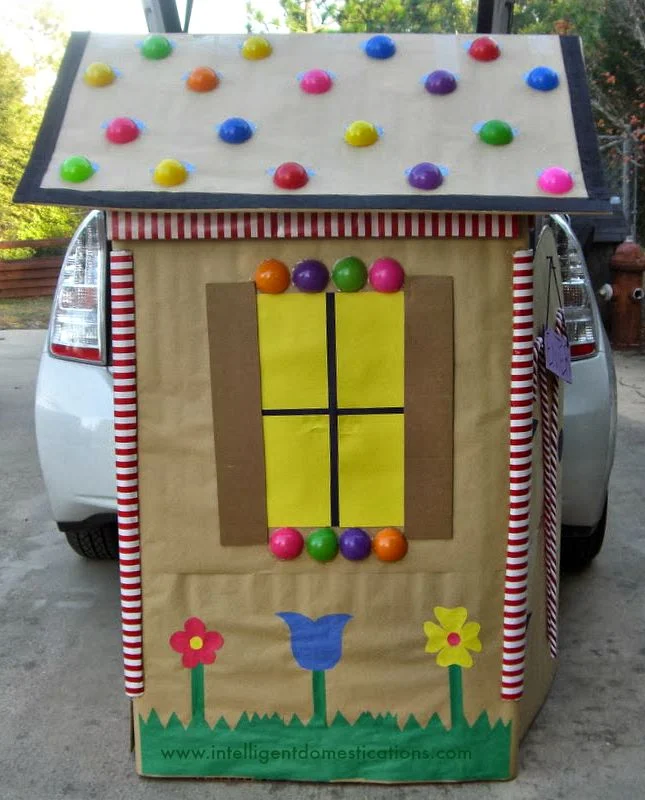 candy cottage made from a cardboard box covered in brown paper with three flowers painted along the bottom, a yellow window made out of paper with brown shutters and colorful dots along the bottom and colorful dots on the roof