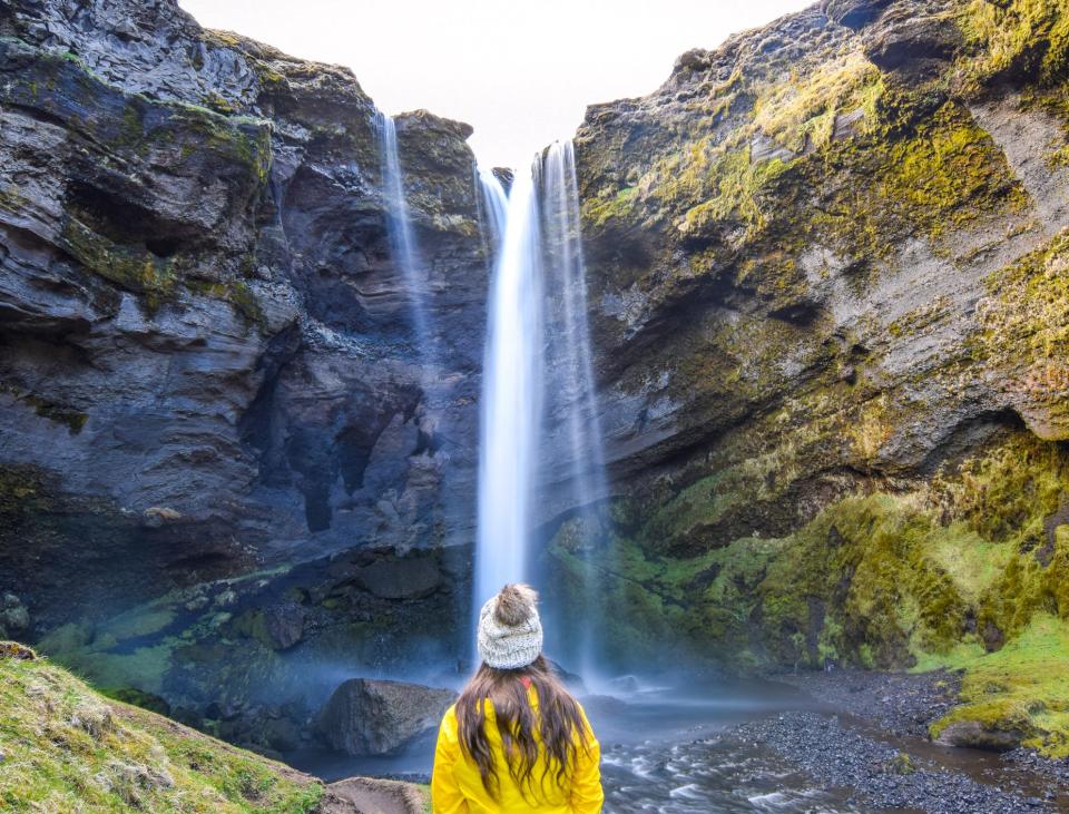 The author in a yellow jacket and gray pom pom hat standing ni front of a waterfall in Iceland