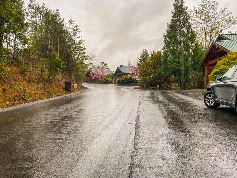 A wet road in the rain with trees on the left and cabins and a parked car on the right. The sky is gray and cloudy.