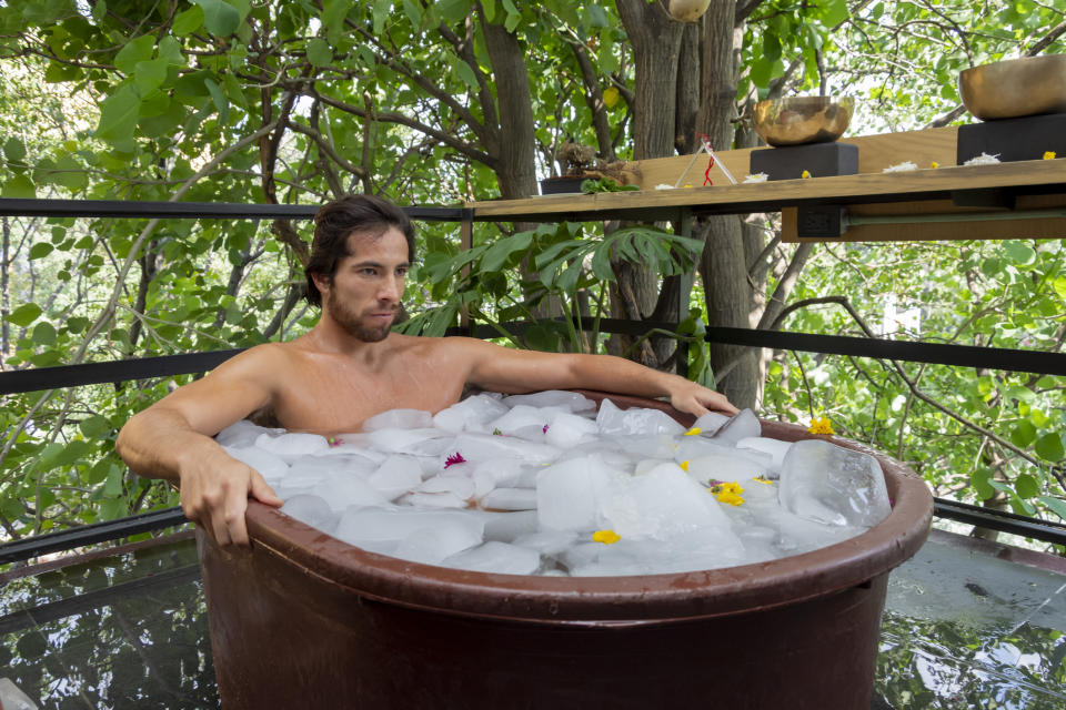 A man is sitting in a wooden tub filled with ice cubes and flower petals. He looks relaxed and contemplative, surrounded by lush greenery