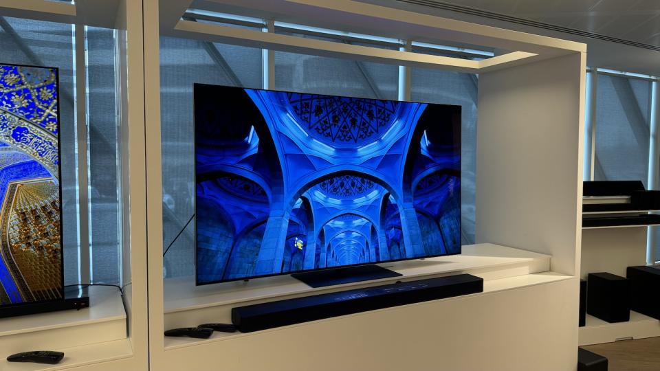 The LG G4 OLED TV photographed on a white stand in a showroom, with a soundbar positioned in front. On the screen is the blue ceiling of a building.