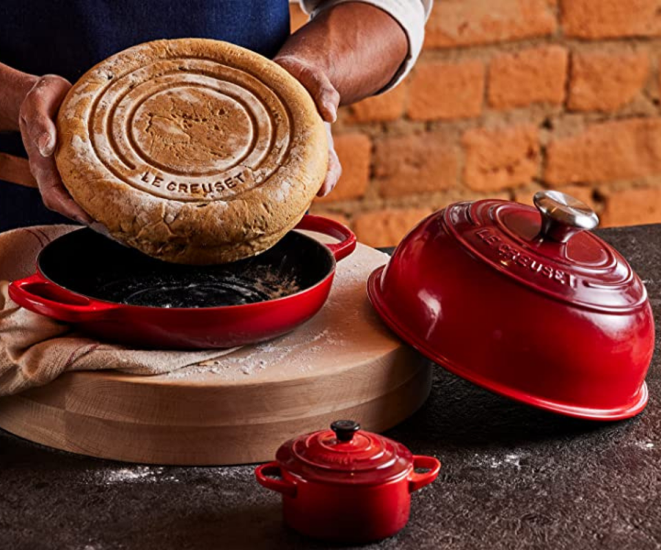 A photo of a man holding a round bread loaf upside down inside a red Le Creuset cast iron bread oven with the lid on the right on a dark surface.