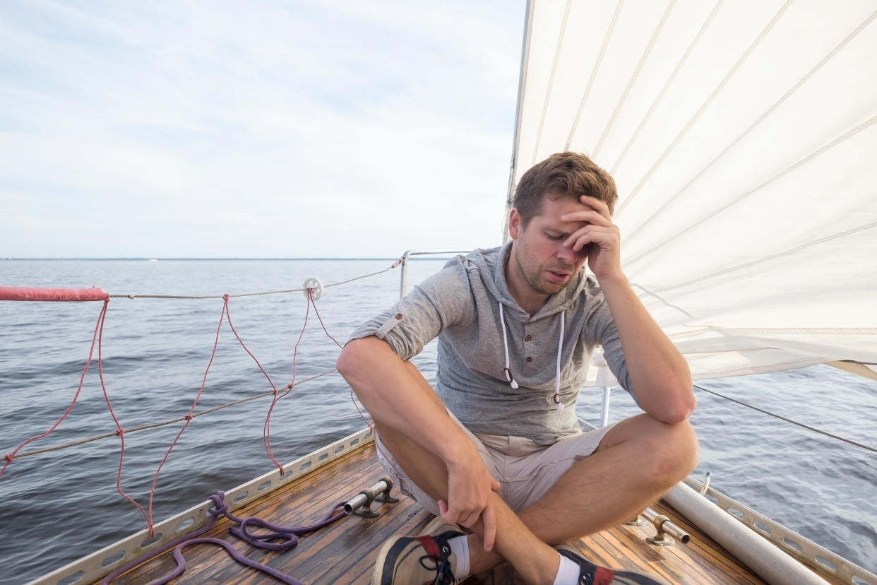 A man sitting on a boat with his eyes closed, holding his hand up to his head and looking distressed.