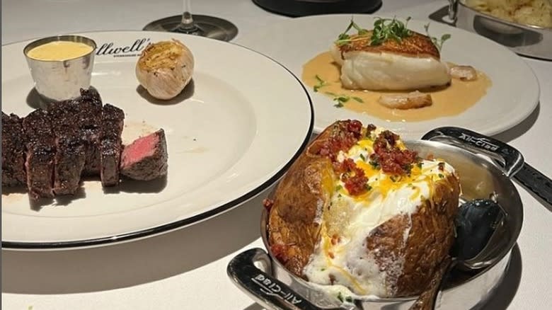 steak on white plate next to baked potato and halibut