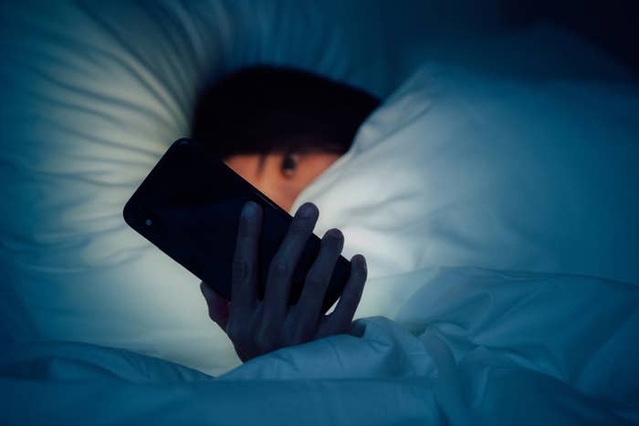 A woman under blanket and holding phone that's illuminating her face in the dark