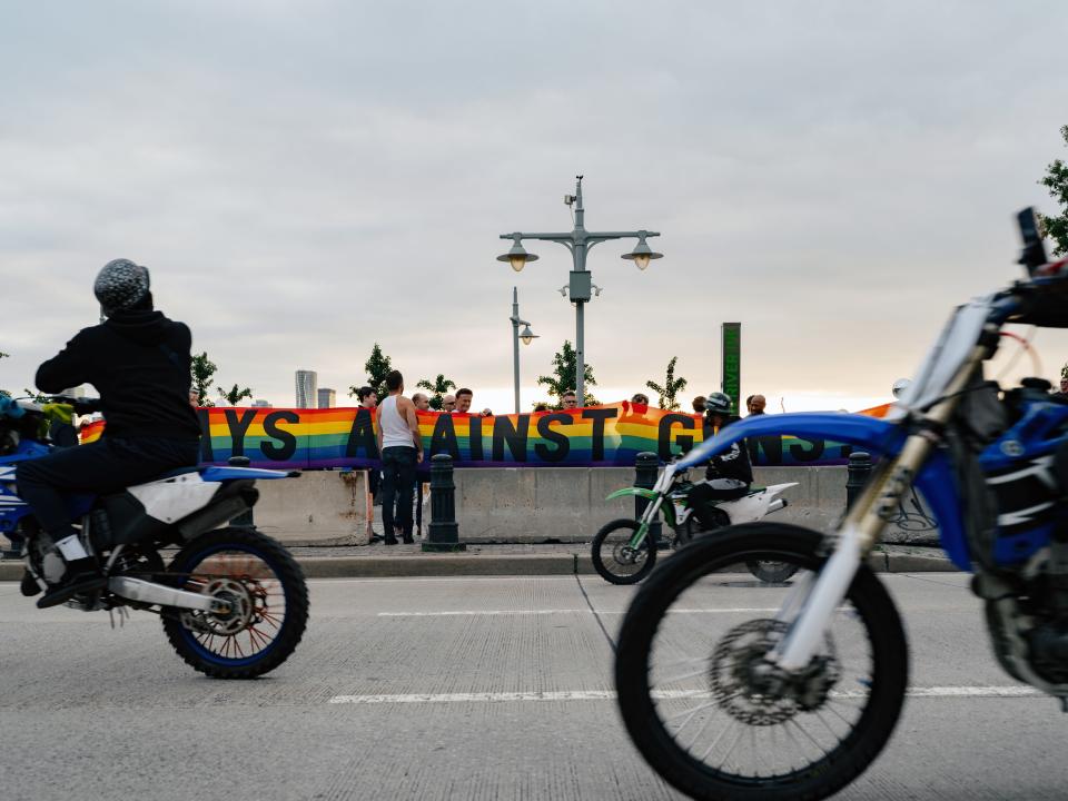 A group of road bikers look at a large rainbow banner reading “GAYS AGAINST GUNS” along the Westside Highway.
