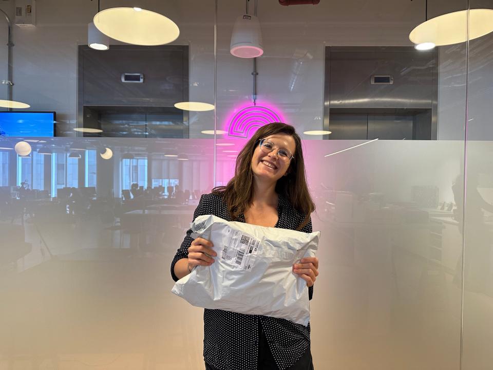 A woman with long brown hair and gold glasses smiling at the camera while holding a white plastic bag in front of her chest.