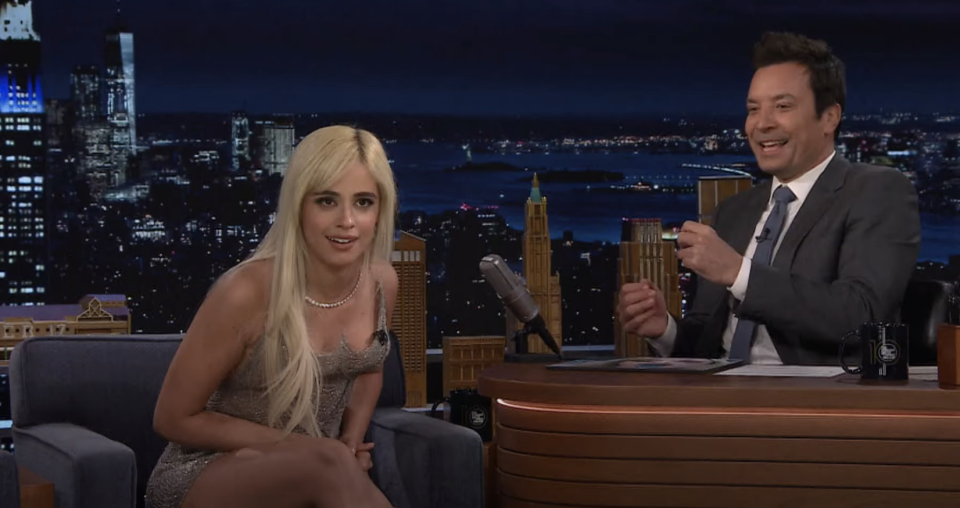 Camila Cabello in a sleeveless, fitted dress, and Jimmy Fallon in a suit, talking and smiling on "The Tonight Show" set with a cityscape background