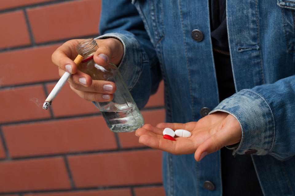 A person wearing a denim jacket holds a cigarette and a glass bottle in one hand and several pills in the other. The image emphasizes substance use