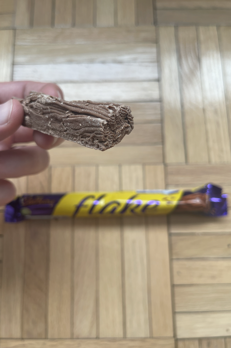 Hand holding a partially unwrapped Cadbury Flake chocolate bar above wooden floor