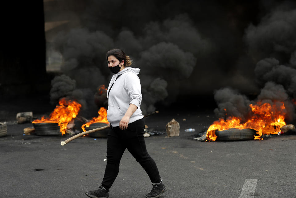 A protester passes in front of burned tires as she blocks a main highway, during a protest in the town of Zouk Mosbeh, north of Beirut, Lebanon, Monday, March 8, 2021. The dayslong protests intensified Monday amid a crash in the local currency, increase of consumer goods prices and political bickering between rival groups that has delayed the formation of a new government. (AP Photo/Hussein Malla)