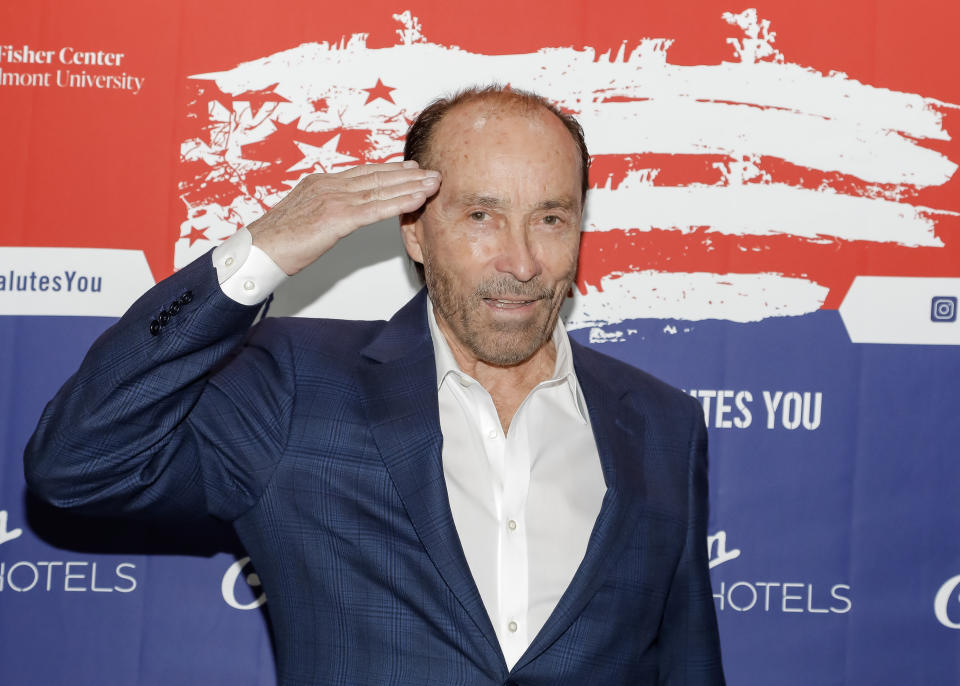 Lee Greenwood salutes on a red carpet, wearing a checked suit jacket and shirt, in front of a patriotic-themed backdrop