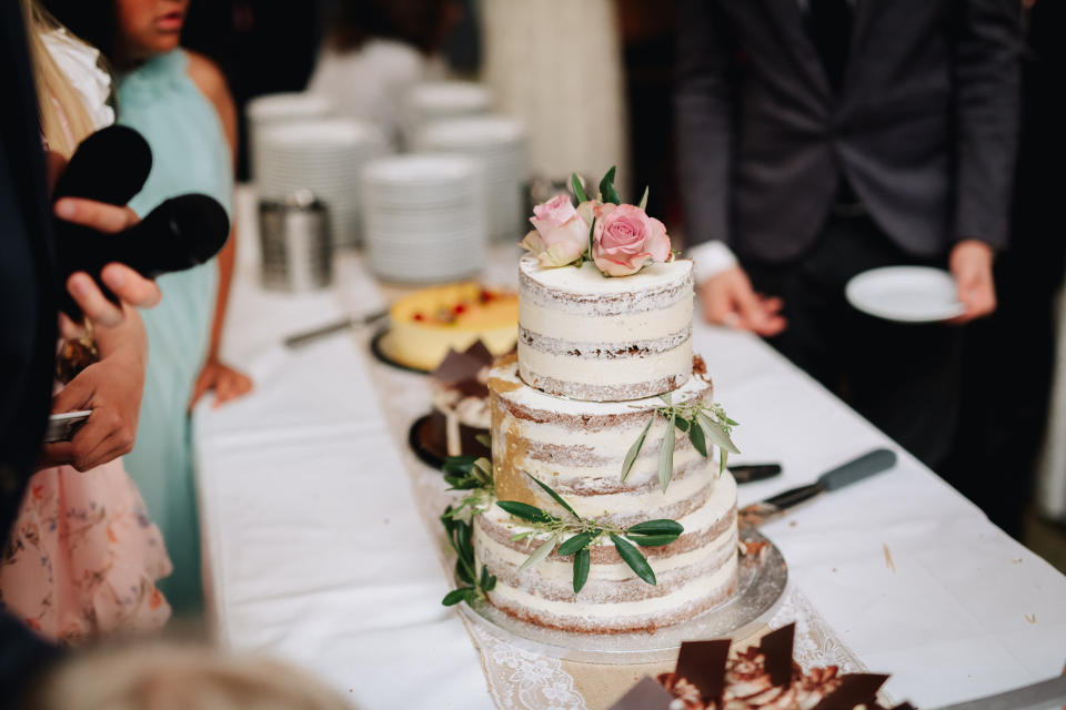 A close-up of a tiered wedding cake with minimal frosting, decorated with pink roses and greenery, surrounded by wedding guests