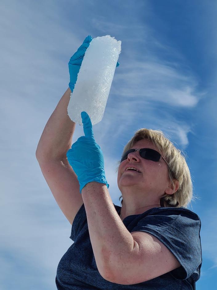 liane benning blonde woman wearing disposable gloves holds up inspects a cylinder of ice against blue sky