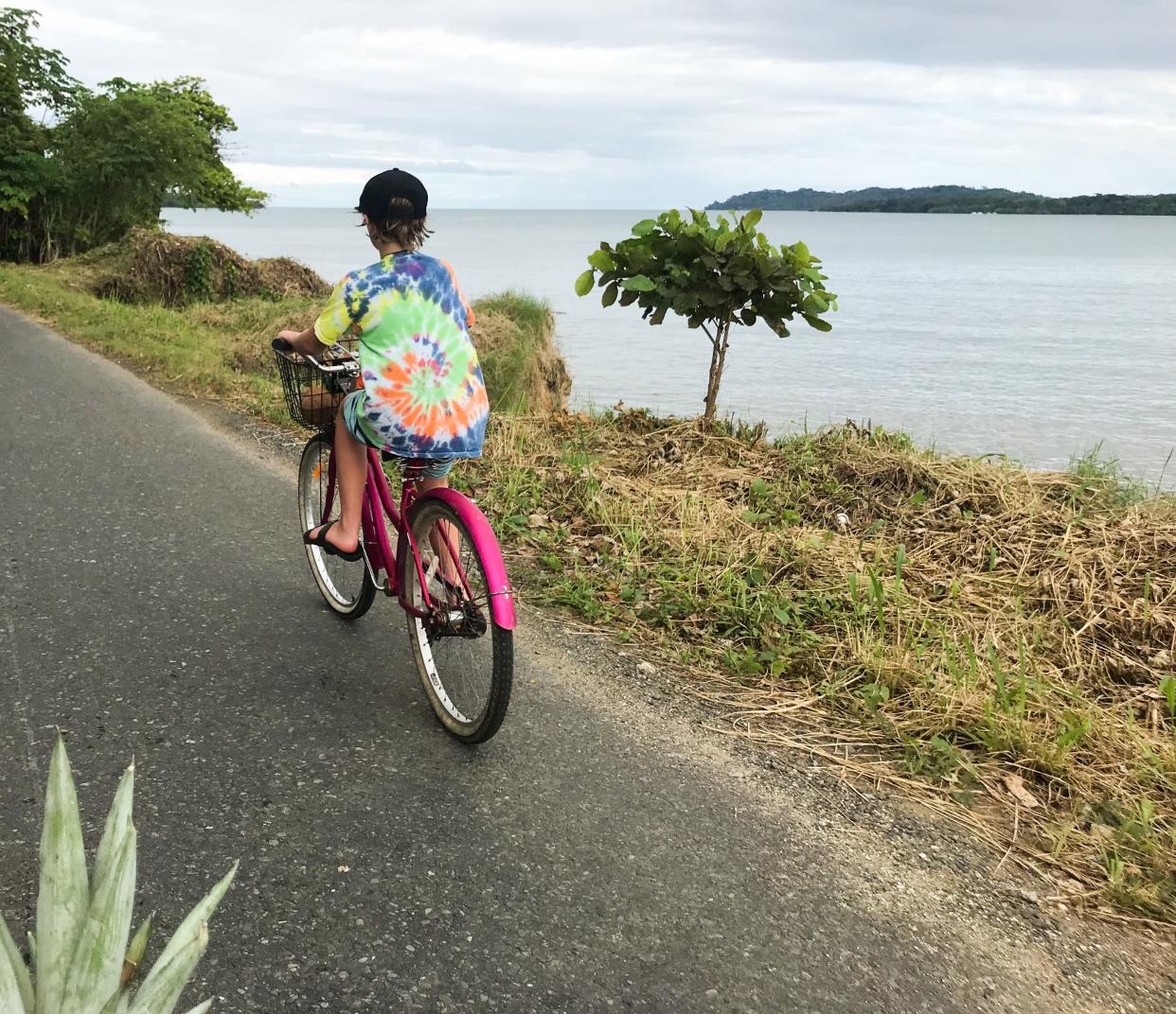 A boy wearing a tie-dye shirt riding a bike near an ocean.