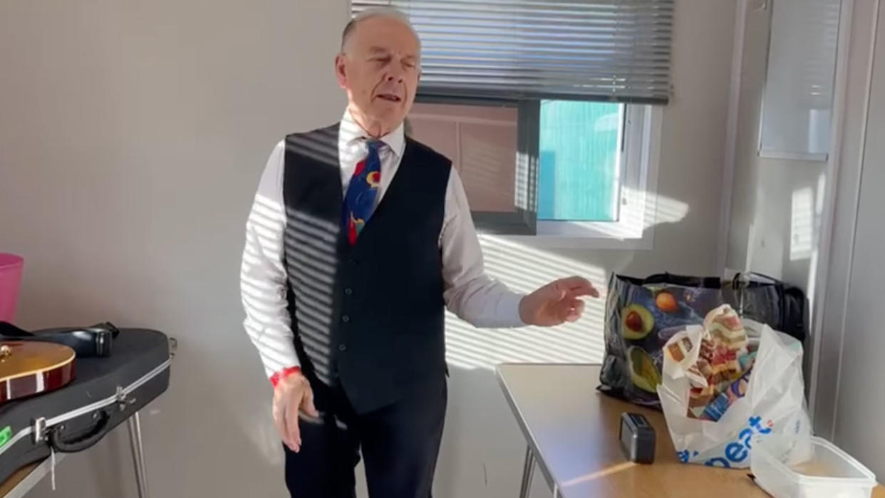  King Crimson's Robert Fripp dressed in a white shirt, colourful tie, dark waistcoat and trousers, with one arm outstretched mid-dance. He's photographed backstage in a small white dressing room with his guitar on a table to the left of the photo and a carrier bag of food to the right of the image 