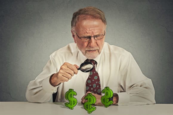 A senior man with a magnifying glass looking at dollar signs on a table.