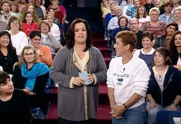 Rosie O'Donnell and a guest stand together smiling in a talk show setting with an audience behind them