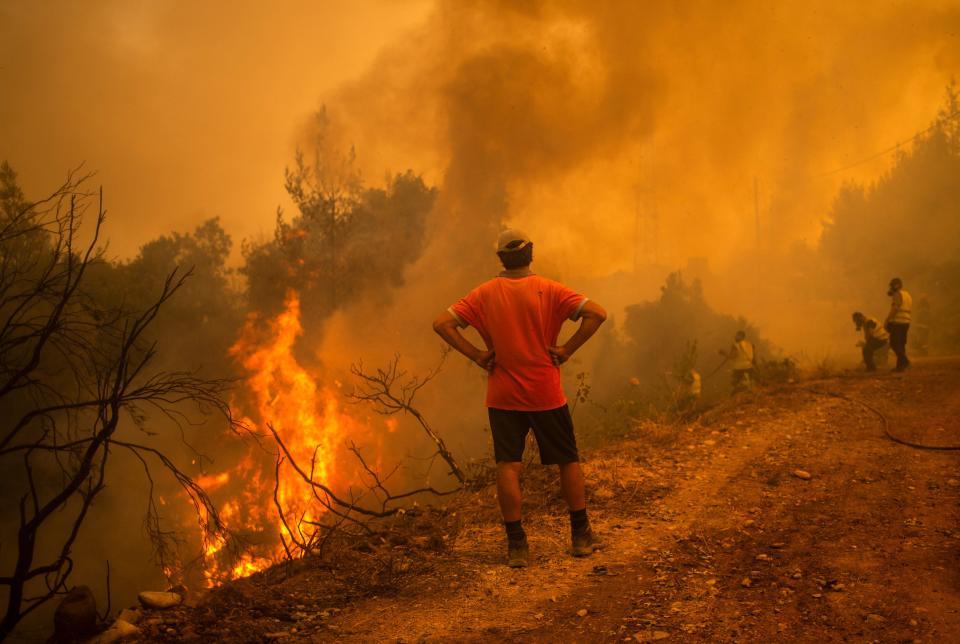 Under a cloud of smoke, a volunteer, with hands on hips, watches firefighters use a water hose near the burning blaze of a forest fire.