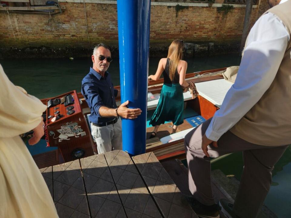 A skipper stands on a speedboat wearing a blue shirt and grey trousers and sunglasses