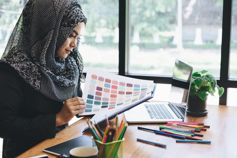 Beautiful young muslim creative designer woman using color palette samples and laptop in office.