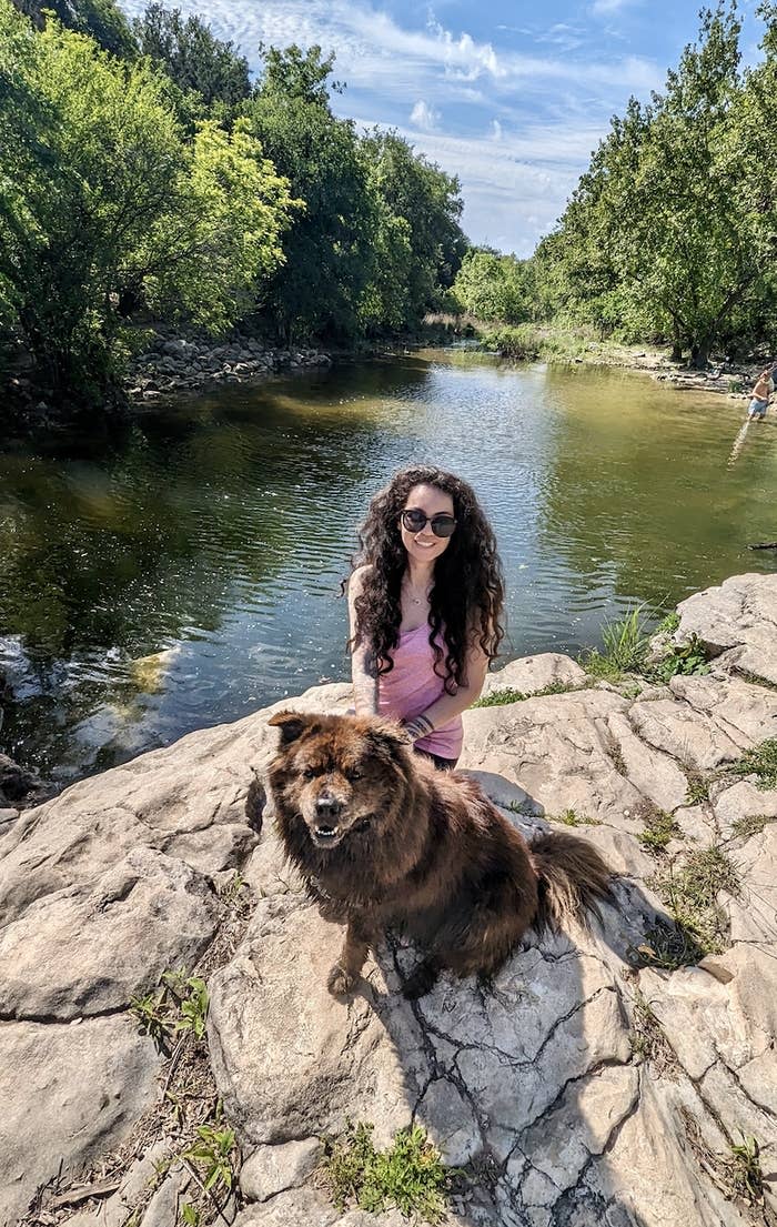 A woman with long curly hair and sunglasses poses on rocky terrain by a serene river with a fluffy brown dog. Greenery surrounds the river in the background