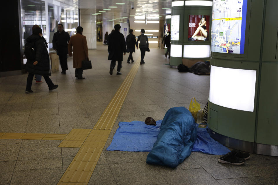 In this Friday, Jan. 10, 2020, photo, commuters walk past a sleeping homeless man at Shinjuku Station, in Tokyo. The dozens of homeless people sleeping rough in shuttered Tokyo subway stations worry that with Japan's image at stake authorities will force them to move ahead of the Olympics. (AP Photo/Jae C. Hong)