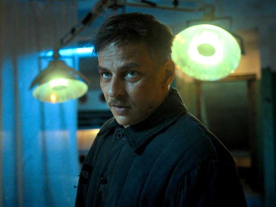 Dimitri (a European man with short brown hair and a mustache also called "Enzo" for a code name) stands in a dark room.