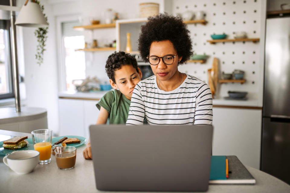 Mid Adult Mother and her Teenage Son Sitting at the Kitchen Counter, Having Breakfast. Mother Trying to Working Online while Her Son Distracting Her, Waiting for a Hug.