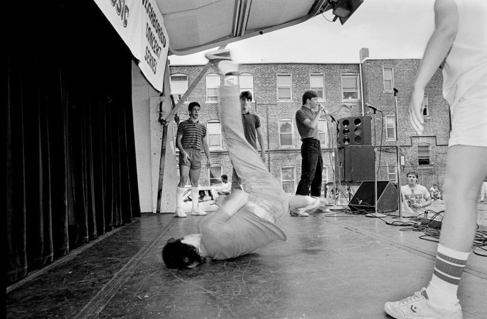 jordan knight breakdances in the center of a stage as danny wood and joey mcintyre watch from the background and donnie wahlberg stands at the front of the stage and raps into a microphone