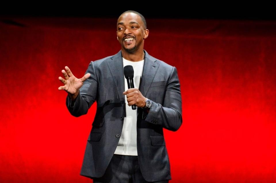 Anthony Mackie in a suit, holding a microphone, speaking warmly to an audience in front of a red background