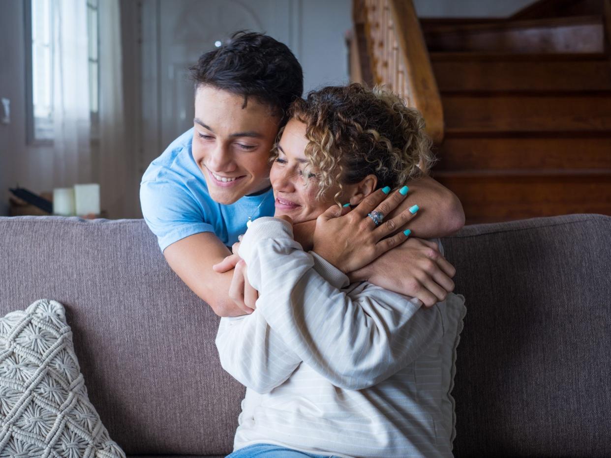 Woman sitting on couch, her teenage son standing behind her smiling and hugging her.