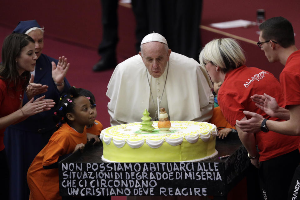 Pope Francis blows a candle atop of a cake he was offered on the eve of his 82nd birthday during audience with children and family from the dispensary of Santa Marta, a Vatican charity that offers special help to mothers and children in need, in the Paul VI hall at the Vatican, Sunday, Dec. 16, 2018. The writing on the bottom of the cake reads: " We can not get use to decay and poverty situation that are around us. A Christian must react ". (AP Photo/Gregorio Borgia)