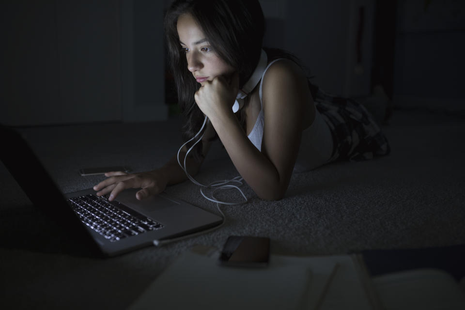 Serious teenage girl using laptop on dark bedroom floor