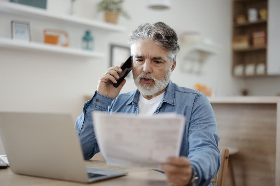 Man reviewing document while talking on phone, sitting at desk with laptop, in a home office setting