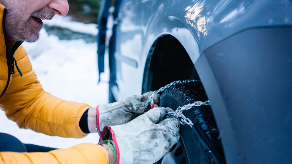 A person puts snow chains on a tire while parked in snowy conditions.  - Westend61/Getty Images