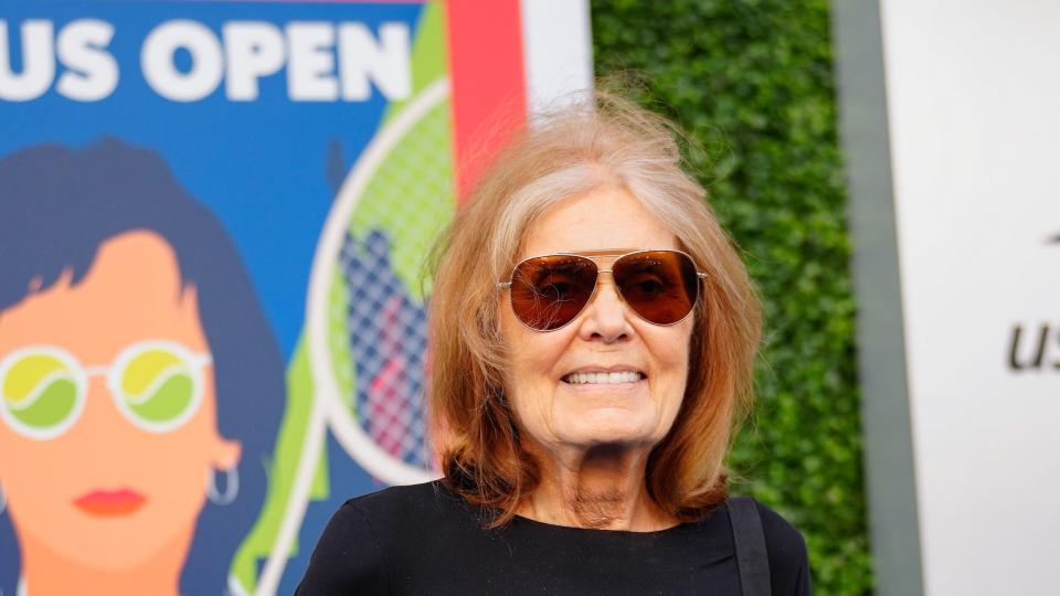 gloria steinem stands in front of a greenery wall with a poster on it, steinem smiles for photos and wears a black shirt and brown aviator sunglasses
