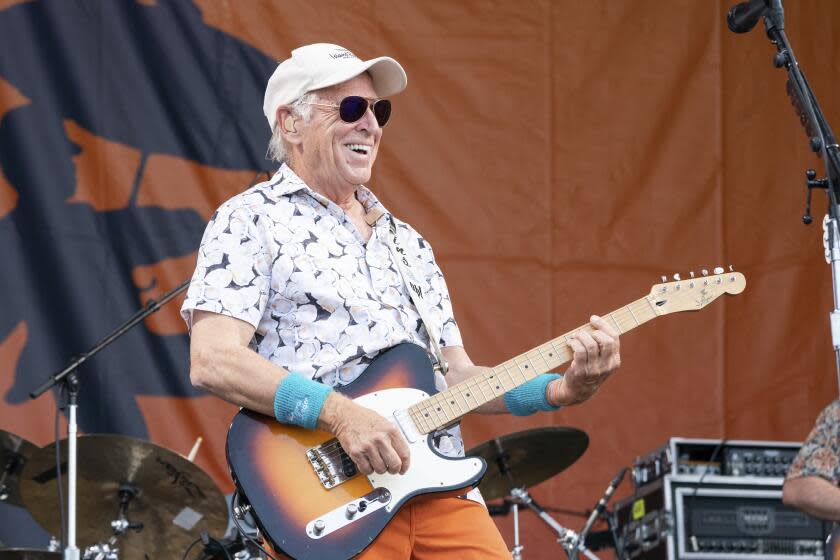 Jimmy Buffett smiles while playing an electric guitar and wearing a beige baseball cap.