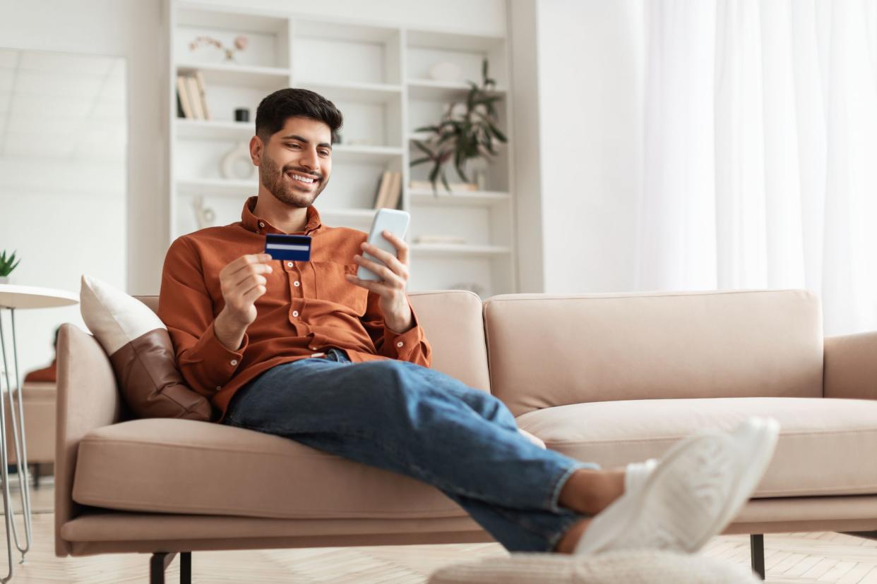 Fast Online Shopping. Smiling young Arab guy holding debit credit card in hand and using cell phone, making financial transaction sitting on the couch at home in living room, free copy space