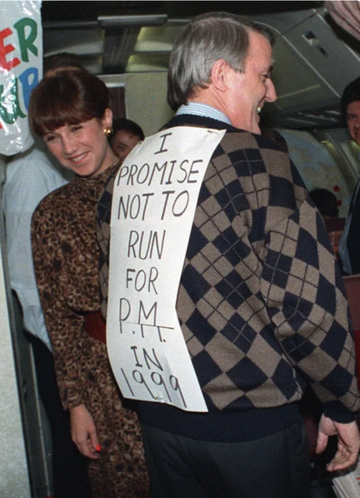 Prime Minister Brian Mulroney, supported by his wife Mila, jokes with the media travelling on his campaign plane Nov. 22, 1988 during the flight home to Ottawa from Baie Comeau. Mulroney wore the sign that said he would not run in 1999, a response to questions earlier in the day regarding his intentions for a third term of office.