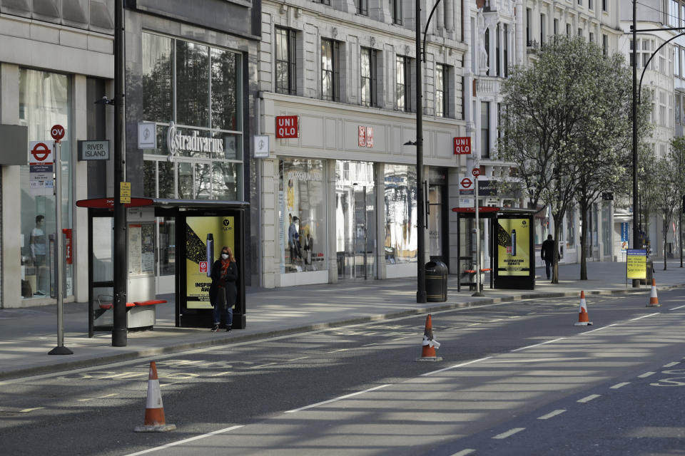 FILE - In this Tuesday, March 24, 2020 file photo, a woman wearing a face mask to protect against coronavirus, waits at a bus stop on a deserted Oxford Street in London. The U.K. economy has officially fallen into a recession after official figures showed it contracting by a record 20.4% in the second quarter as a result of lockdown measures put in place to counter the coronavirus pandemic. The slump recorded by the Office for National Statistics follows a 2.2% quarterly contraction in the first three months of the year. (AP Photo/Matt Dunham, File)