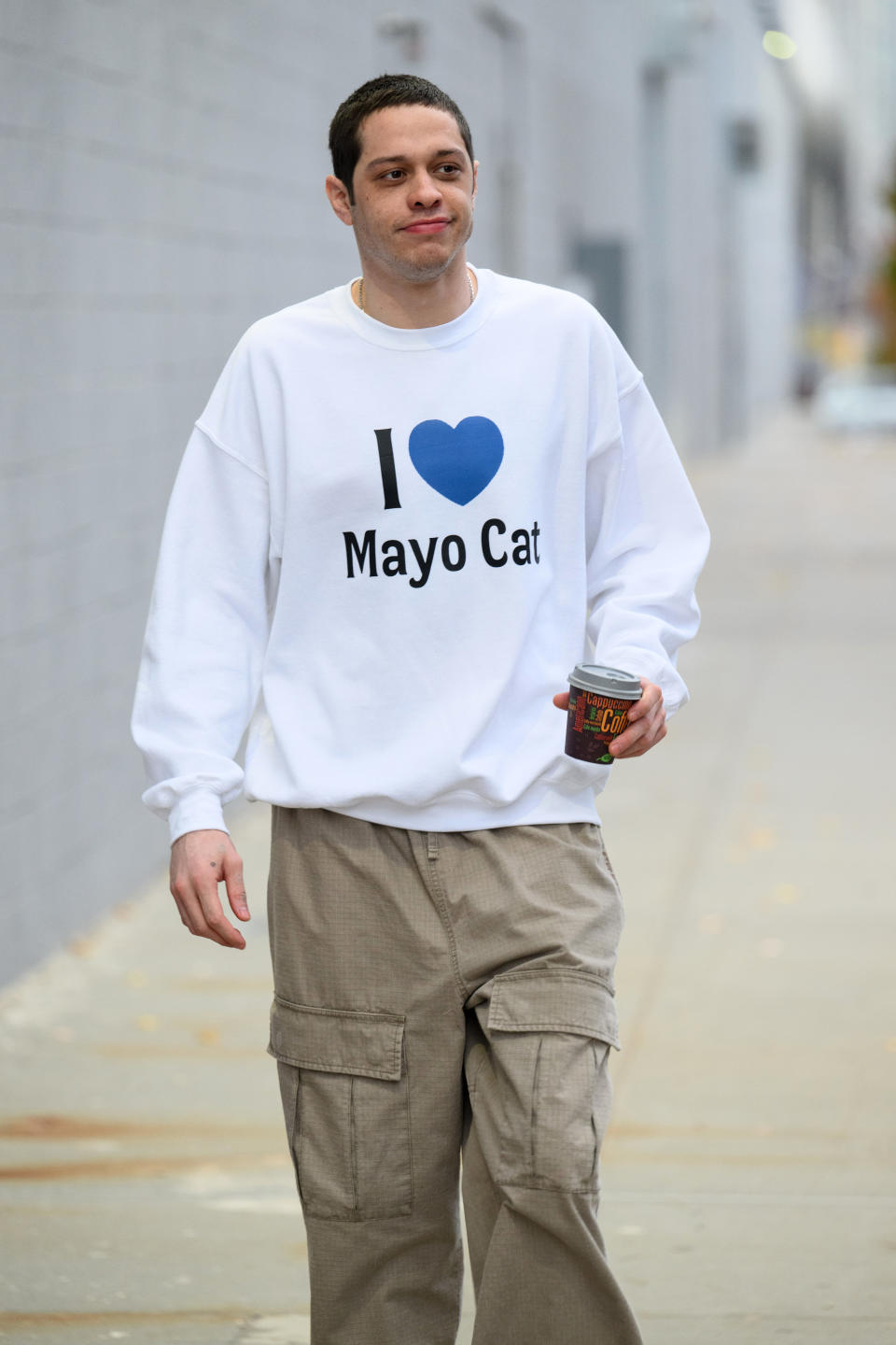 Pete Davidson walking outside in casual cargo pants, holding a cup and wearing a sweatshirt with the text "I [heart] Mayo Cat."