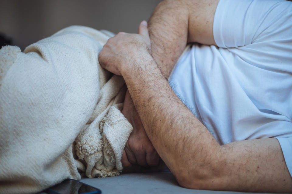 A person with folded arms lies on a bed, holding a blanket close. Only the arms and torso in a white shirt are visible. Image conveys resting or comfort