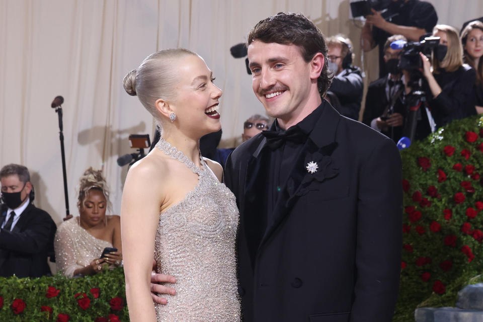 Phoebe Bridgers, in a glittering halter-neck dress, and Paul Mescal, in a black tuxedo, smiling together at a formal event, surrounded by press and attendees