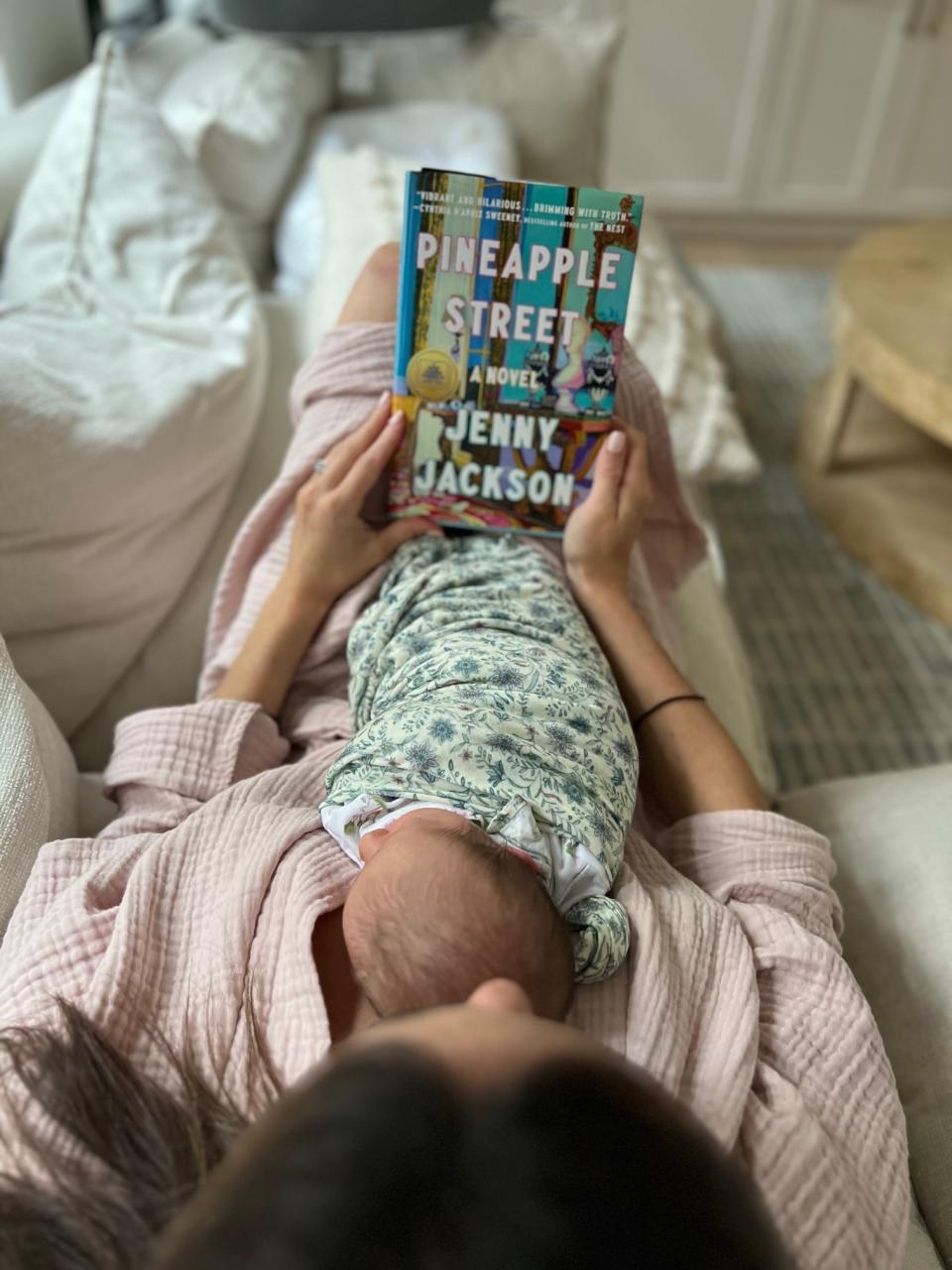 Hannah lying on a couch and reading a book with the baby resting on her chest