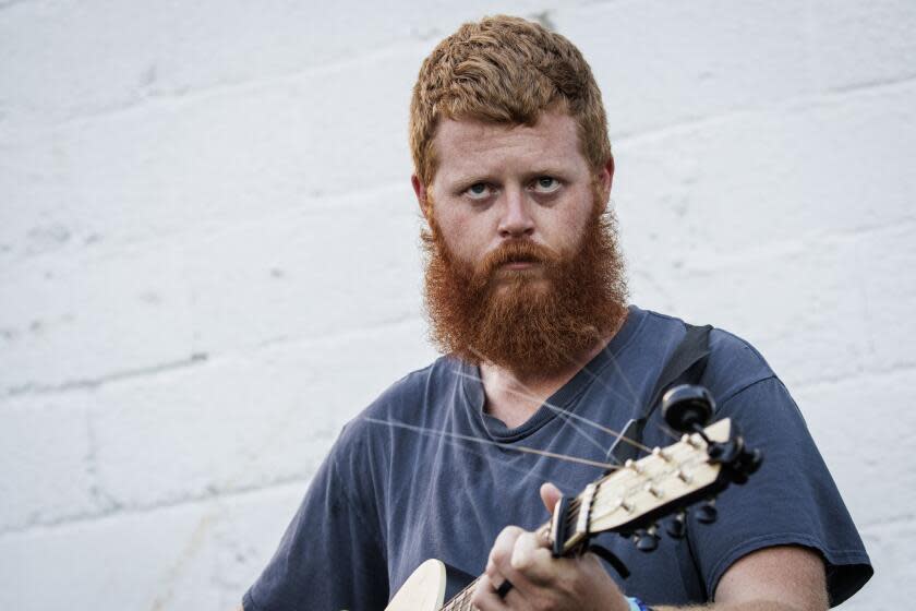 Oliver Anthony, with red hair and beard, is staring intensely as he plays his guitar while wearing a blue t shirt