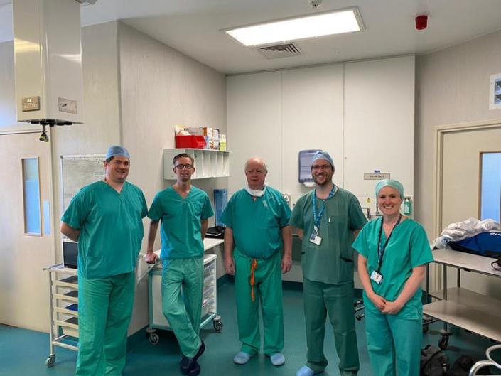 Five people stand in an operating theatre wearing medical scrubs.